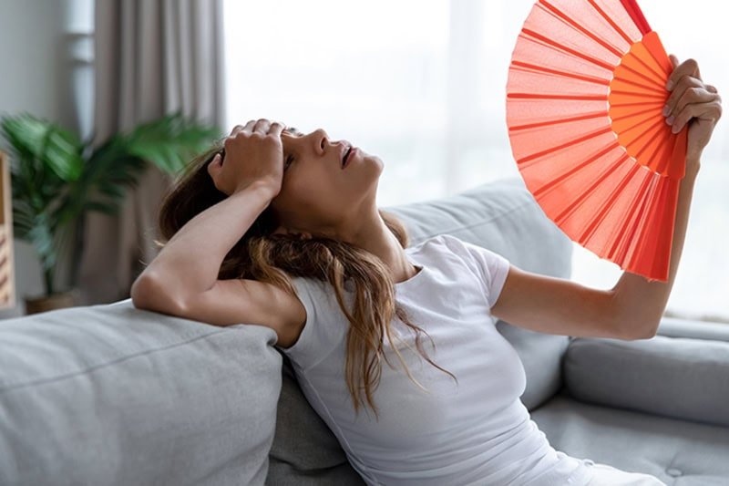 AC Blowing Hot Air. Side view exhausted young biracial girl using paper waver, suffering from hot summer weather or high temperature at home. Unhappy overheated millennial mixed race woman feeling uncomfortable indoors.