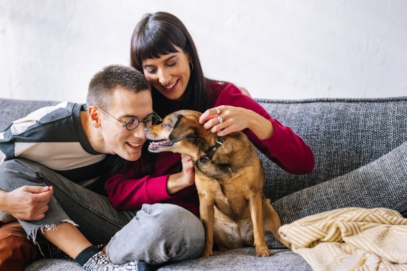 A lovely couple sitting on the sofa and playing with their dog. Everybody is smiling.