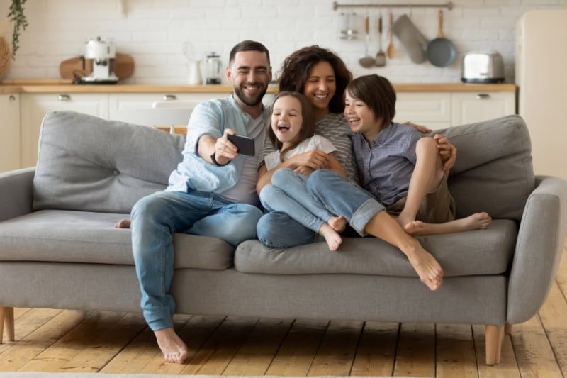 Smiling parents with little kids laughing using smartphone together sitting on couch at home. Happy father holding phone taking selfie with children. Family watching video having fun with cellphone.